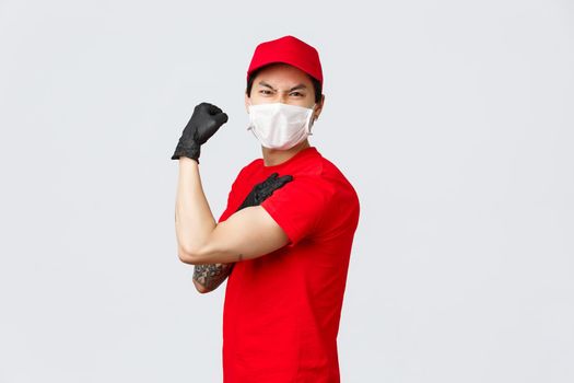 Strong asian guy show-off his strength, carry large parcels at work. Delivery man in uniform flex biceps and grimacing, wear medical mask and gloves, advertisement of carrier and order delivery.