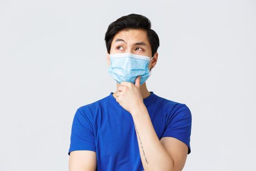 Different emotions, lifestyle and leisure during coronavirus, covid-19 concept. Close-up of thoughtful asian man facing difficult decision, wear medical mask touch chin and look left, thinking.