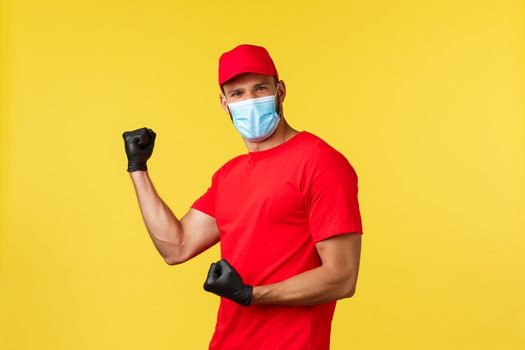 Express delivery during pandemic, covid-19, safe shipping, online shopping concept. Enthusiastic and energized courier in red uniform and medical mask, say yes fist pump, winning, yellow background.