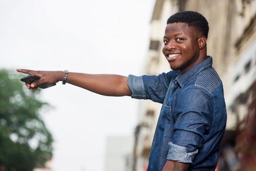 Young man smiling with his hand raised, holding a mobile phone and standing in the street waiting for taxi.