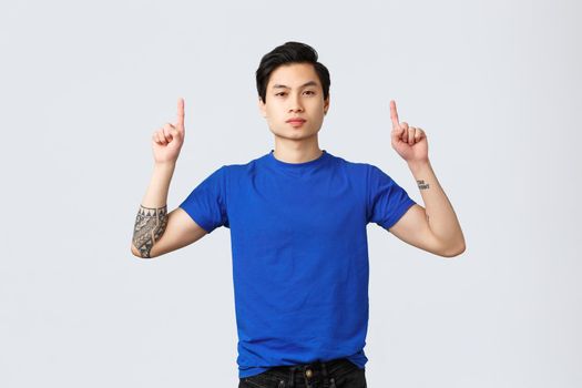 Different emotions, people lifestyle and advertising concept. Confident, serious asian man in blue t-shirt with tattoos, pointing fingers up, inform clients, showing way to banner, grey background.