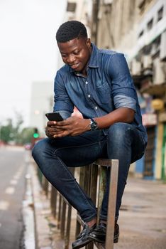 Portrait of a smiling young man sitting with a mobile phone on an iron bar in the street