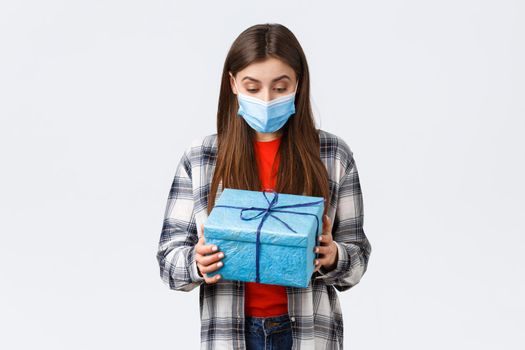 Covid-19, lifestyle, holidays and celebration concept. Happy and surprised birthday girl, employee receive present from coworkers, looking at wrapped gift with amazed grateful face in medical mask.