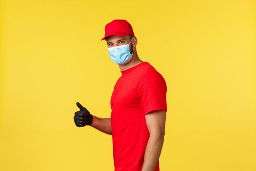 Express delivery during pandemic, covid-19, safe shipping, online shopping concept. Smiling handsome courier in red uniform and medical mask, show thumb-up, recommend package deliver service.