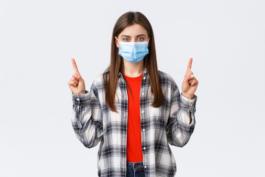 Coronavirus outbreak, leisure on quarantine, social distancing and emotions concept. Confident and determined young woman self-isolating home, pointing fingers up, wear medical mask.