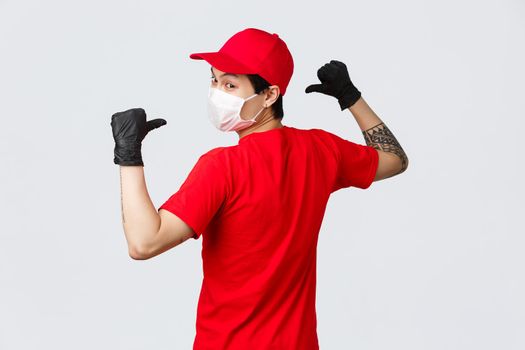 Back view of asian delivery man in medical mask and protective gloves, wear red cap, t-shirt, turn to camera, pointing back to show company logo on uniform, logistics, courier and shopping concept.