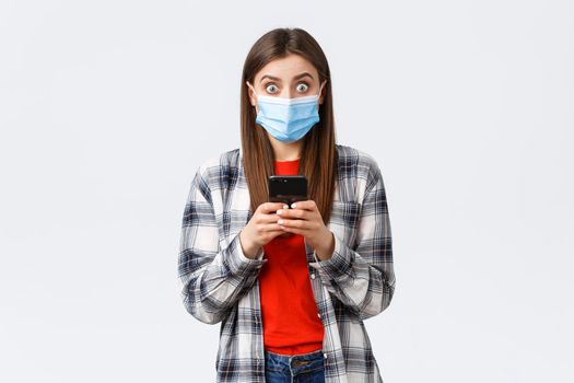 Different emotions, covid-19, social distancing and technology concept. Surprised young woman got news via text message, raising head from mobile phone and stare camera impressed, wear medical mask.