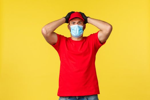 Express delivery during pandemic, covid-19, safe shipping, online shopping concept. Nervous and concerned courier in red uniform, medical mask, staring shocked, feel trouble, grab head.