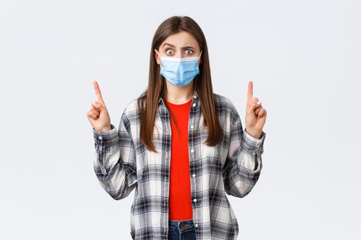Coronavirus outbreak, leisure on quarantine, social distancing and emotions concept. Concerned and puzzled young woman in medical mask pointing fingers up at strange, confusing promo.