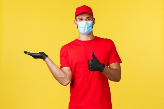 Express delivery during pandemic, covid-19, safe shipping, online shopping concept. Smiling courier in red uniform, medical mask and gloves, showing product and thumb-up in approval.