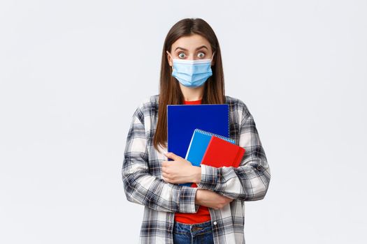 Coronavirus pandemic, covid-19 education, and back to school concept. Surprised female student in medical mask look amazed, widen eyes from big news, carry notebooks, white background.