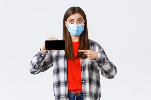 Different emotions, covid-19, social distancing and technology concept. Impressed and surprised girl in medical mask stare astonished, pointing finger at mobile phone screen.