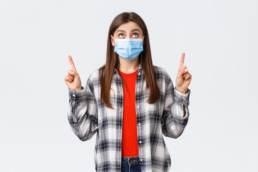 Coronavirus outbreak, leisure on quarantine, social distancing and emotions concept. Surprised and interested cute caucasian girl in casual outfit and medical mask, reading sign upwards, point up.