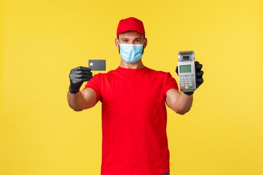 Food delivery, tracking orders, covid-19 and self-quarantine concept. Cheerful young courier in medical mask and gloves, suggest contactless payment with POS terminal and credit card.