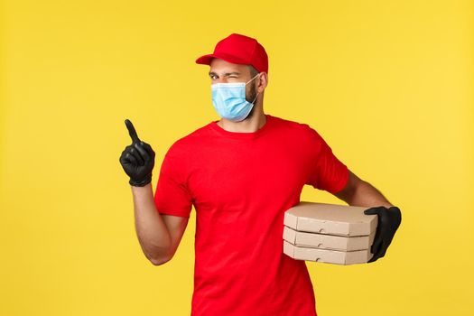 Food delivery, tracking orders, covid-19 and self-quarantine concept. Handsome enthusiastic courier in red uniform and medical mask, wink, pointing upper left corner and hold pizza, click here.