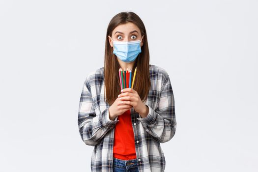 Social distancing, leisure and hobbies on covid-19 outbreak, coronavirus concept. Enthusiastic cute girl in medical mask trying new thing on self-quarantine, showing colored pencils, learn how draw.