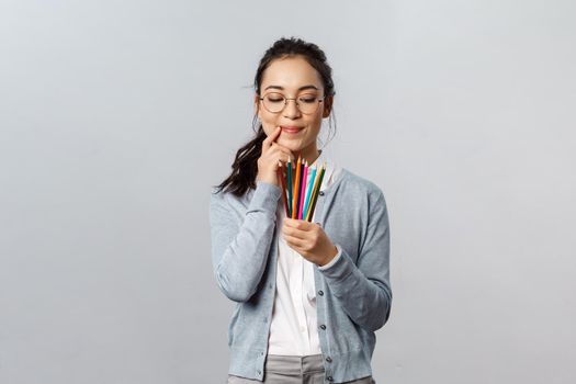 Hobbies, creativity and art concept. Thoughtful young creative female artist touch lip thinking and smiling as look at colored pencils, plan what to draw next for inktober, stand grey background.