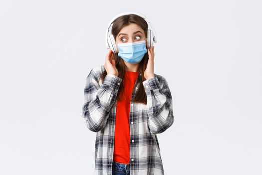Social distancing, leisure and lifestyle on covid-19 outbreak, coronavirus concept. Cute silly caucasian girl in medical mask, listening music in headphones, look left intrigued or surprised.