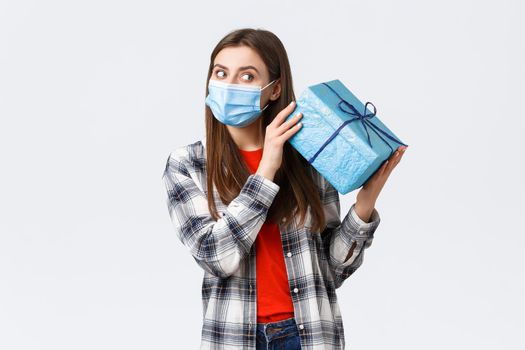 Covid-19, lifestyle, holidays and celebration concept. Cute birthday girl in medical mask, shaking gift box to guess what inside, receive b-day present, celebrating during coronavirus self-quarantine.