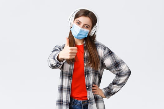 Social distancing, leisure and lifestyle on covid-19 outbreak, coronavirus concept. Pleased cheerful cute girl in medical mask and headphones, showing thumb-up, recommend or approve.