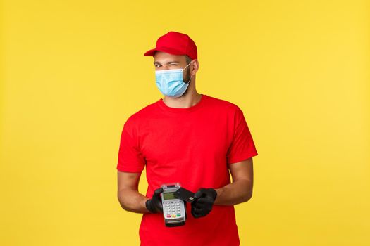 Food delivery, tracking, covid-19 and self-quarantine concept. Friendly courier in red uniform, medical mask and gloves, wink at camera, asking pay contactless with payment terminal and credit card.