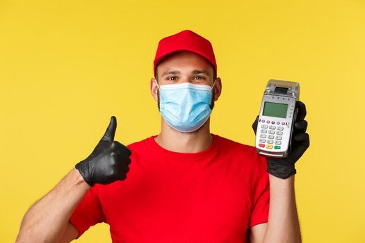 Covid-19, delivery orders, shopping, contactless payment and social distancing concept. Satisfied handsome courier in red uniform, advice use POS terminal for safety during coronavirus pandemic.