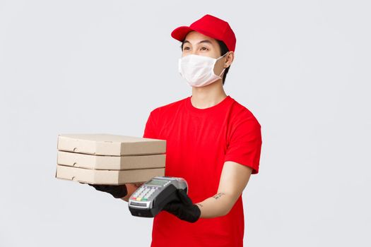 Contactless delivery, safe purchase and shopping during coronavirus concept. Friendly courier in red uniform cap and t-shirt, giving client pizza delivery order and payment terminal, grey background.