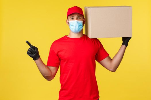 Express delivery during pandemic, covid-19, safe shipping, online shopping concept. Upset and sad courier in red uniform and medical mask, pointing finger left, hold box package on shoulder.