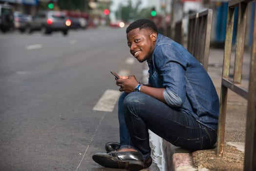 young man sitting in jeans near iron bar with mobile phone in hand looks at camera smiling.