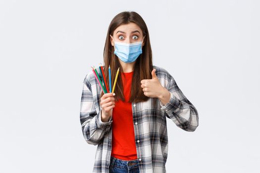 Social distancing, leisure and hobbies on covid-19 outbreak, coronavirus concept. Excited and impressed girl in medical mask recommend cool new online lessons, learn draw, show thumb-up and pencils.