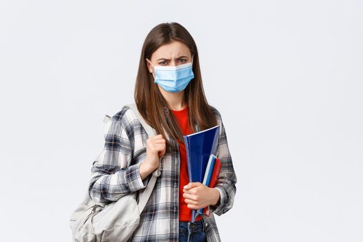 Coronavirus pandemic, covid-19 education, and back to school concept. Upset disappointed female student in medical mask hate studying in university during infection oubreak, frowning condemn dean.