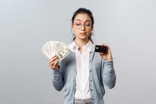 Business, finance and investment concept. Thoughtful indecisive cute asian girl look tempting at cash dollars, holding money and credit card, shopping, think where invest, grey background.