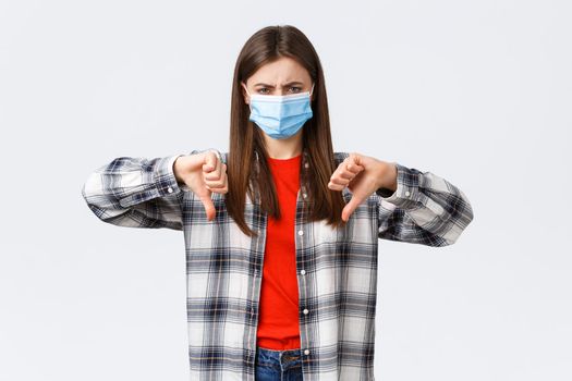 Coronavirus outbreak, leisure on quarantine, social distancing and emotions concept. Disappointed and mad young woman in medical mask vote against, show thumb-down and grimacing dismay.