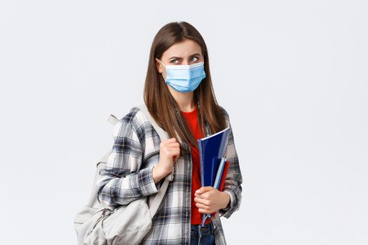 Coronavirus pandemic, covid-19 education, and back to school concept. Displeased female student in medical mask and backpack, look behind at upper left corner with disdain or dismay, hold notebooks.