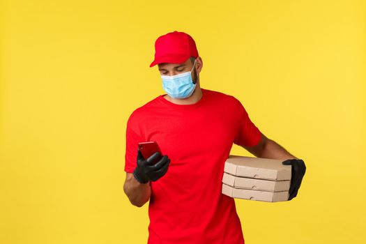 Food delivery, tracking orders, covid-19 and self-quarantine concept. Courier checking client adress on phone as bring pizza order, calling customer, standing yellow background.