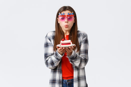 People lifestyle, holidays and celebration, emotions concept. Cute tender young b-day girl in glasses, blowing lit candle on birthday cake, making wish and celebrating at party, white background.