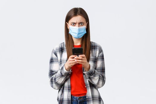 Different emotions, covid-19, social distancing and technology concept. Frustated and confused young woman in medical mask react to strange message, hold mobile phone, frowning camera.