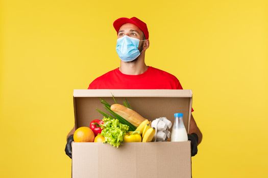 Takeaway delivery, covid-19 quarantine and groceries concept. Young courier in red cap and t-shirt uniform, medical mask, look up thoughtful, searching customer apartment, hold box with food order.