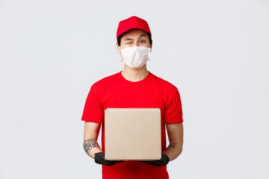 Friendly asian delivery man in red cap and t-shirt, wear protective gloves and medical mask for client and employees safety, house delivery during covid-19 quarantine, holdin box package.