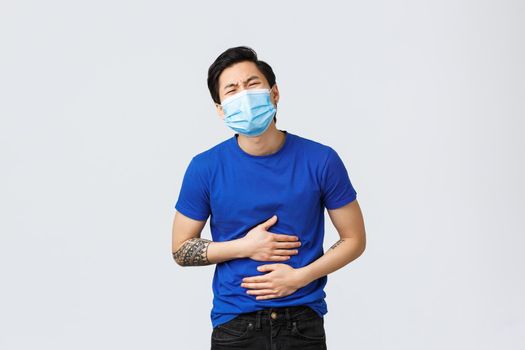 Different emotions, social distancing, self-quarantine and lifestyle concept. Asian guy suffering huge pain in stomach. Man with stomachache touching belly and bending from ache, food poison.