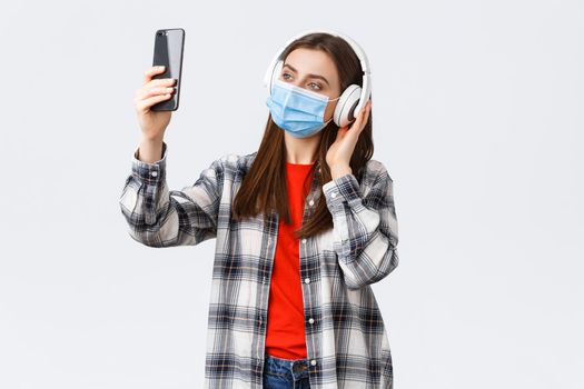 Social distancing, leisure and lifestyle on covid-19 outbreak, coronavirus concept. Woman in headphones and medical mask listening music, taking selfie on mobile phone using filters.