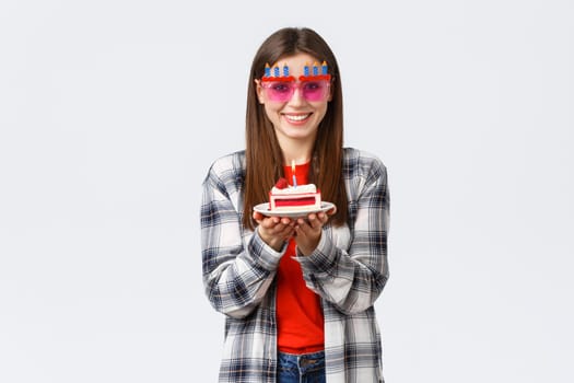 People lifestyle, holidays and celebration, emotions concept. Cheerful cute girl in glasses holding birthday cake, celebrate b-day, making wish to blow lit candle, white background.