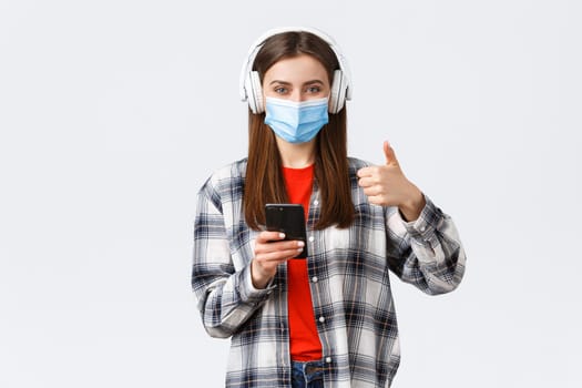 Social distancing, lifestyle on covid-19 outbreak, coronavirus concept. Cheerful pleased good-looking woman in medical mask, approve nice sound of wireless headphones, thumb-up, hold phone.