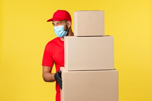 Express delivery during pandemic, covid-19, safe shipping, shopping concept. Friendly handsome courier in red uniform, medical mask, standing near pile of box orders, taking care customers packages.