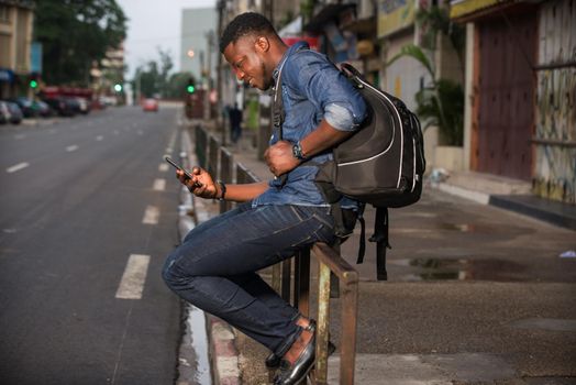 young man sitting in jeans on iron bar looking at mobile phone while smiling.