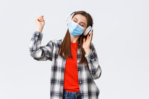 Social distancing, leisure and lifestyle on covid-19 outbreak, coronavirus concept. Carefree tender young woman carried away listening music in headphones, dancing with closed eyes in medical mask.