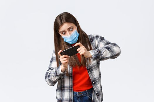 Different emotions, covid-19 pandemic, coronavirus self-quarantine and social distancing concept. Focused and entertained woman in medical mask playing mobile game, tilting body, difficult level.