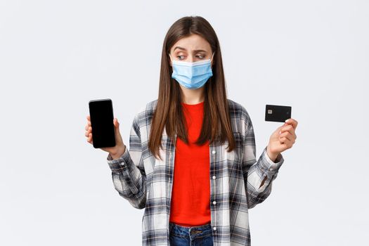 Coronavirus outbreak, working from home, online shopping and contactless payment concept. Unsure girl in medical mask look as making decision at mobile phone, show credit card and smartphone.