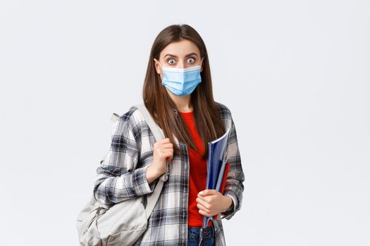 Coronavirus pandemic, covid-19 education, and back to school concept. Shocked and surprised girl in medical mask, student gasping over big news in campus, hold notebooks and backpack.