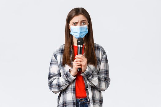 Coronavirus outbreak, leisure on quarantine, social distancing and emotions concept. Confused woman in medical mask look smth strange, holding microphone, perform, white background.